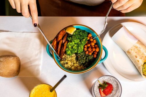 A vibrant vegan meal with fresh ingredients like broccoli, chickpeas, quinoa, served alongside a burrito and refreshing drinks.