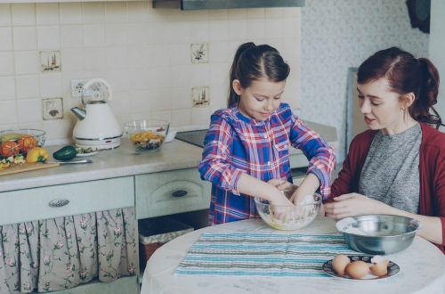 Little cheerful and funny girl helping her mother in the kitchen cropping egg into bowl and mixing dough for cookies. Family, food, home and people concept